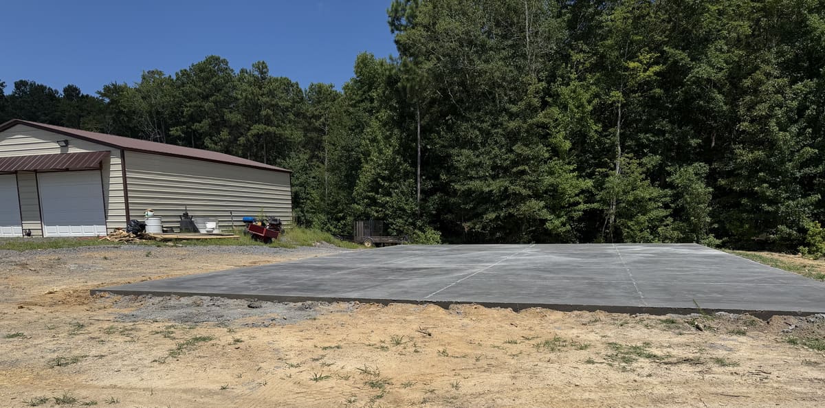 Large cured pad with control joints beside a tan metal shop building with roll-up doors, woods behind