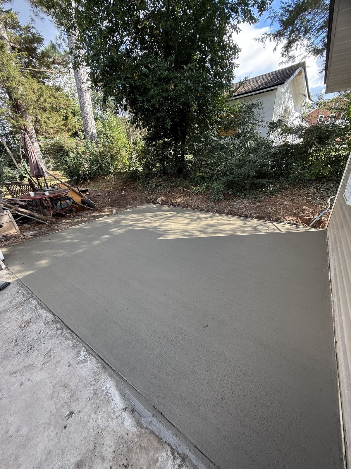 Fresh broom-finished patio slab beside a vinyl-sided house, shaded by tall trees