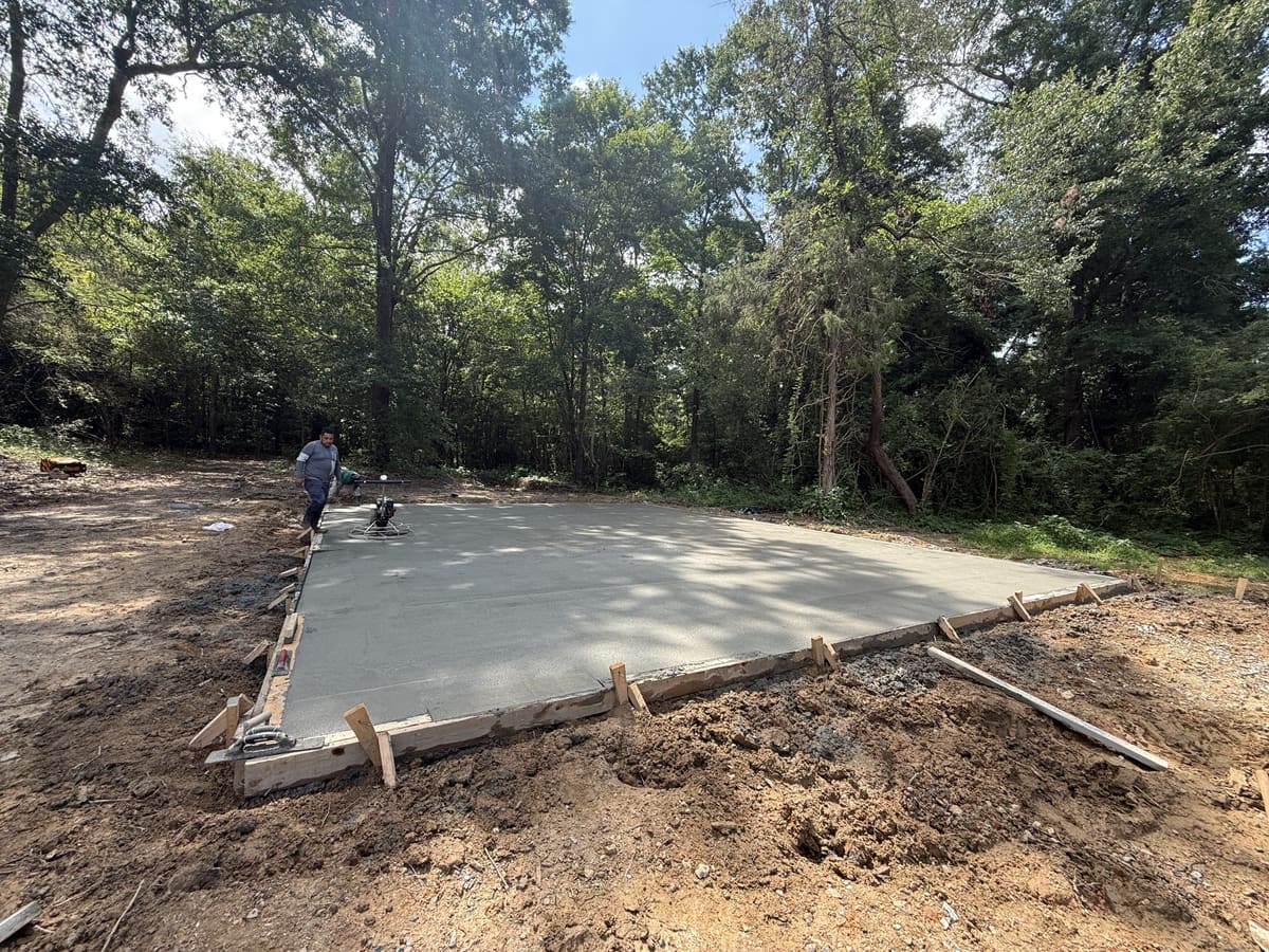 Crew member walking the form edge beside a walk-behind power trowel on a fresh pad in wood forms, hardwoods all around