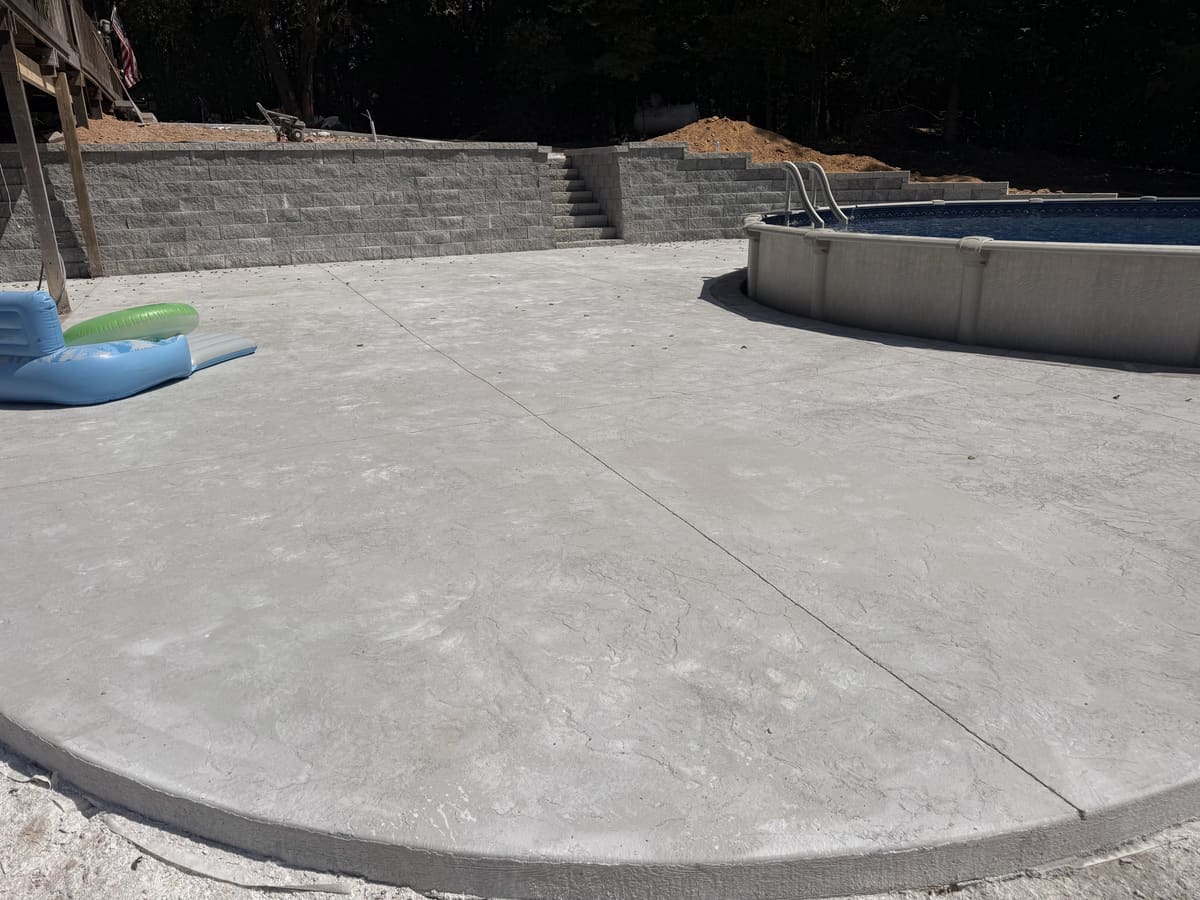 Round pool deck in a seamless stone-texture stamped finish around an above-ground pool, a block retaining wall behind